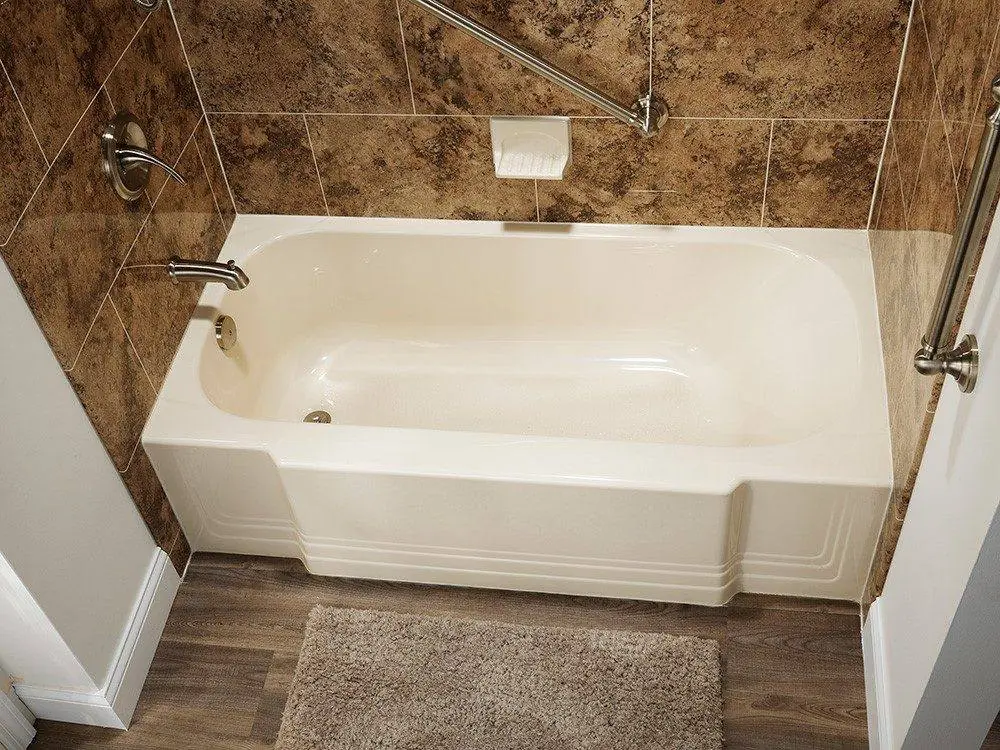 White bathtub with a brown granite look wall surround, a grab bar and a wood look floor