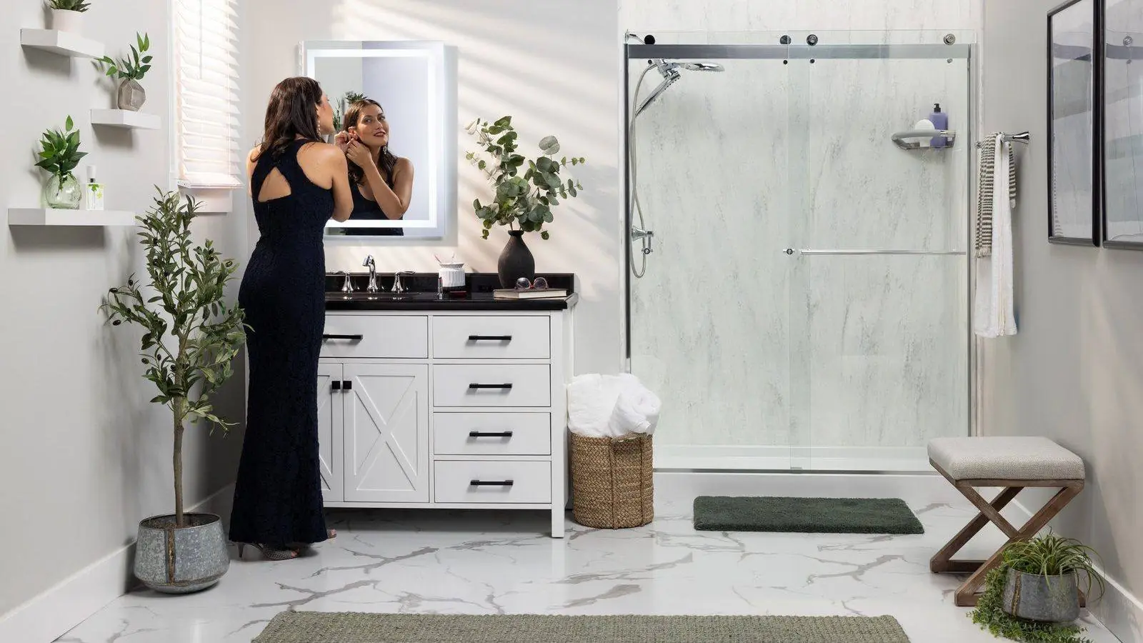 Woman at a lit vanity mirror beside a marble look shower with a sliding glass door